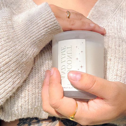 Person holding a candle with a visible brand name, wearing a light sweater and gold ring.