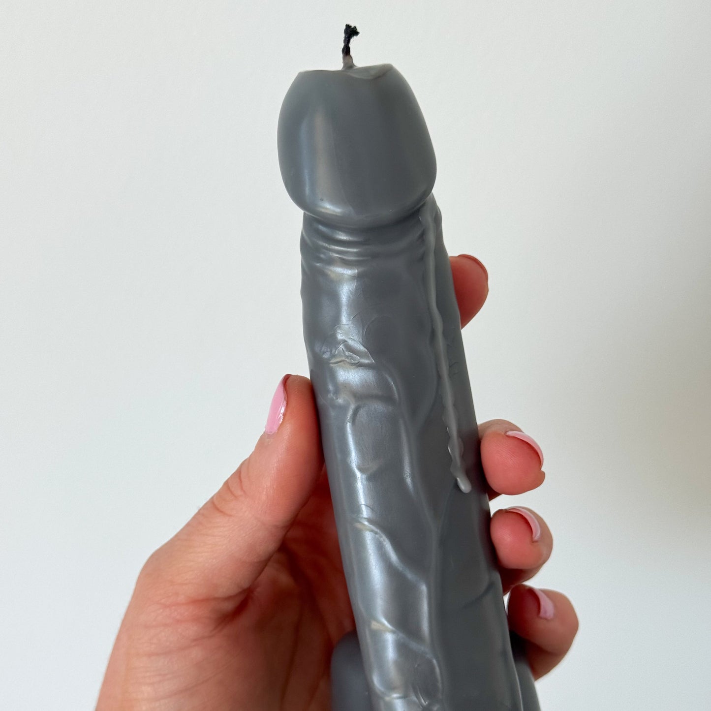 A hand holding a novelty sculptural candle shaped like a stylized penis, made of matte gray wax with a small black wick, against a plain off-white background.