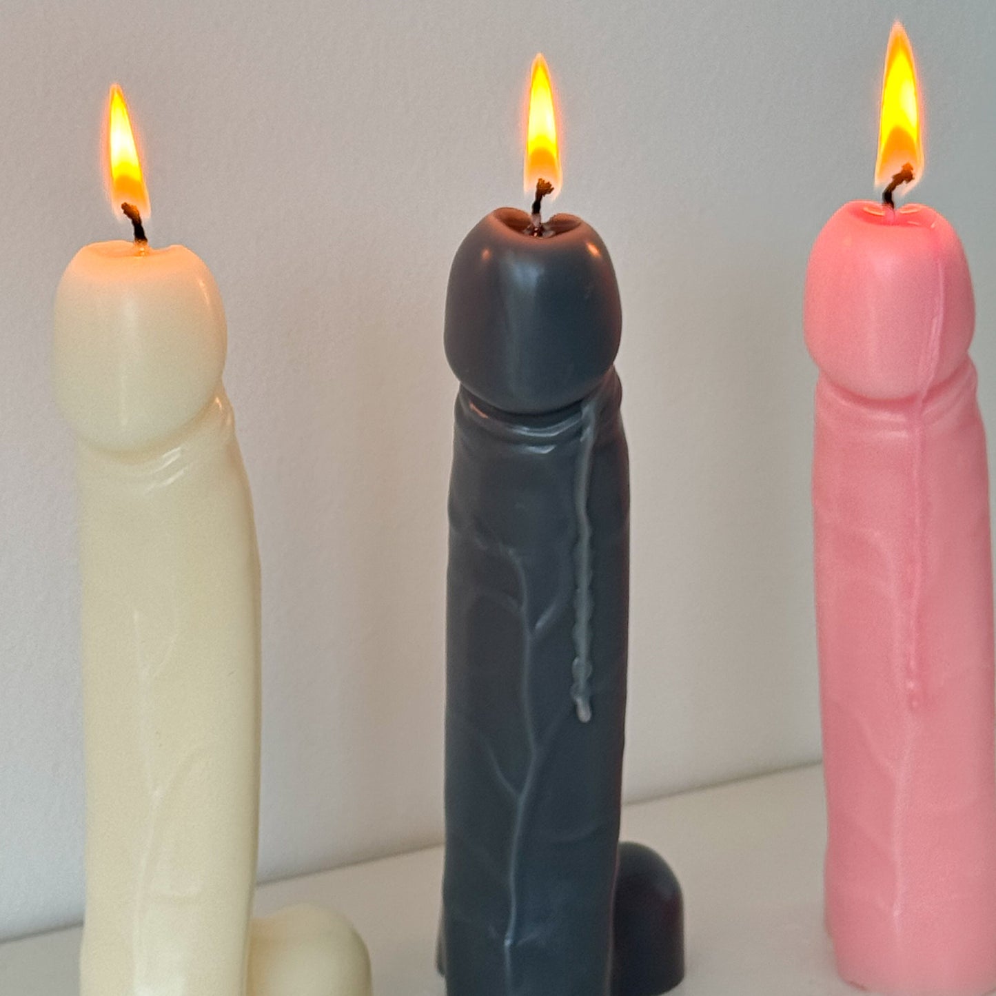 Three novelty pillar candles shaped like male genitalia, shown lit on a white surface against a plain light gray wall. The left candle is cream, the center candle is dark gray, and the right candle is pink.
