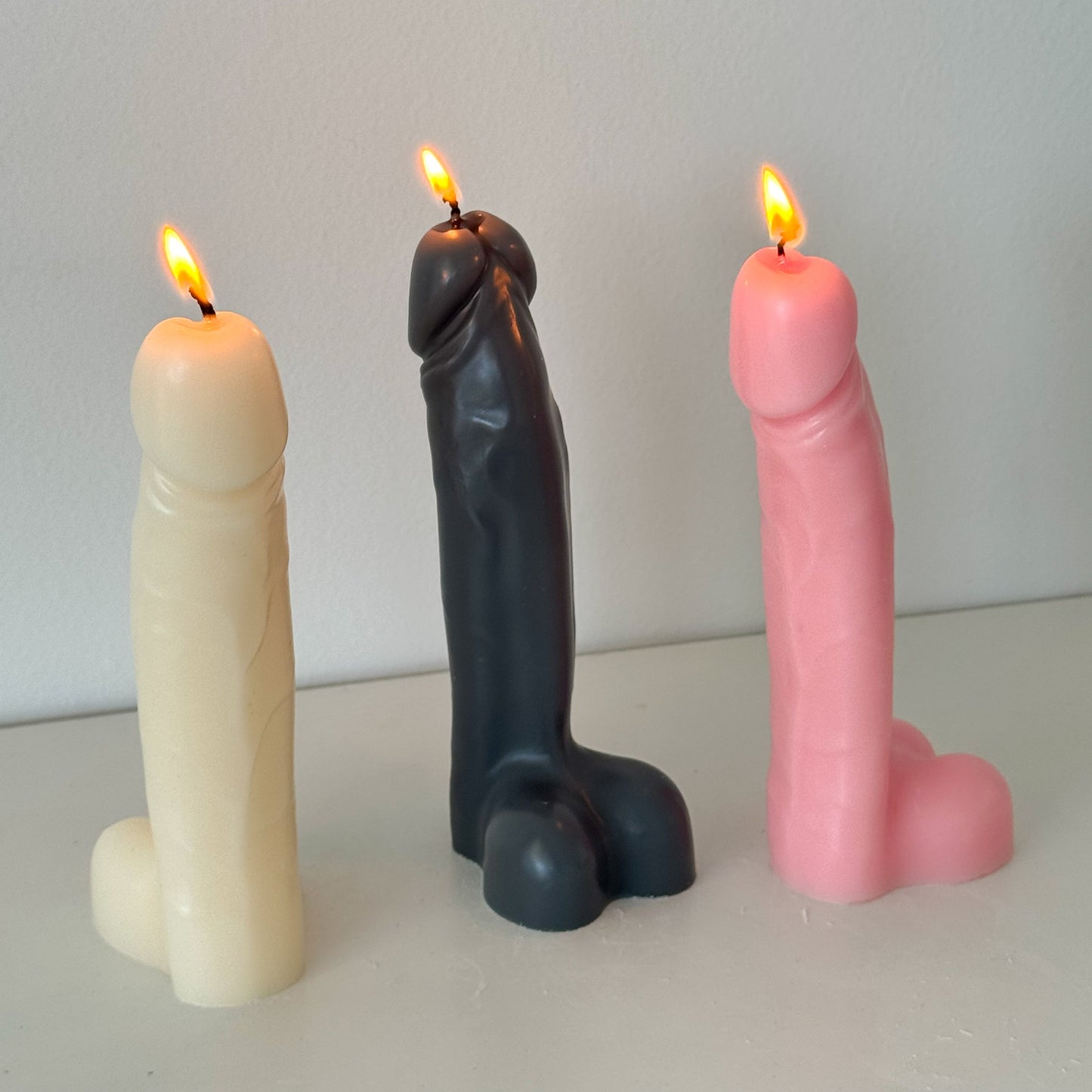 Three novelty sculptural candles shaped like stylized penis forms, standing upright and lit with visible flames. One candle is cream/off-white, one is dark charcoal/black, and one is light pink, set against a plain light gray wall and surface.
