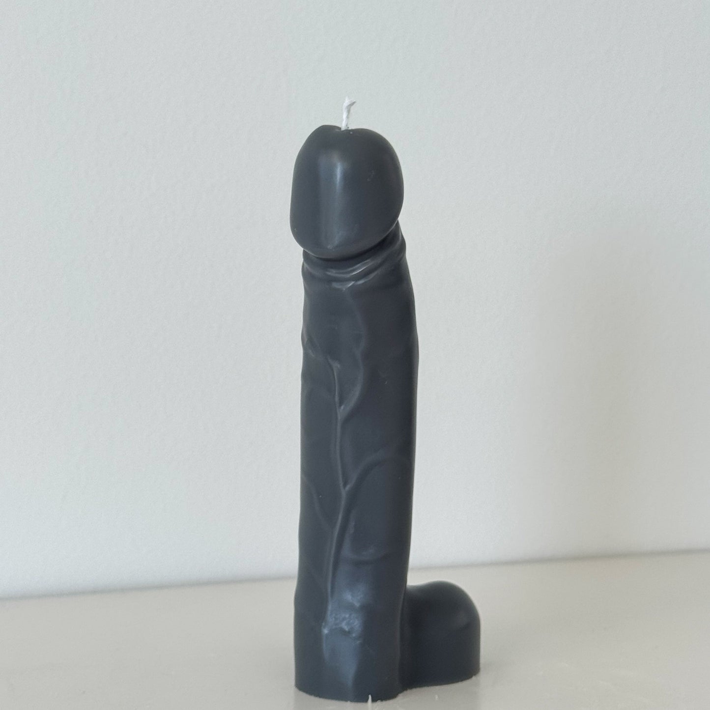 A novelty black silicone candle shaped like a stylized penis, standing upright on a light surface against a plain white wall.