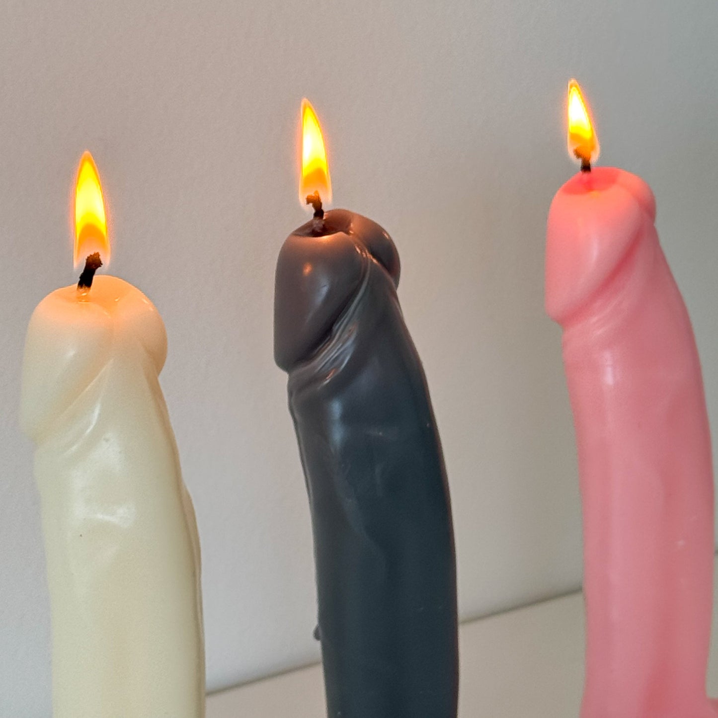 Three novelty penis-shaped candles with lit flames, shown in cream, dark gray, and pink colors against a plain light background.