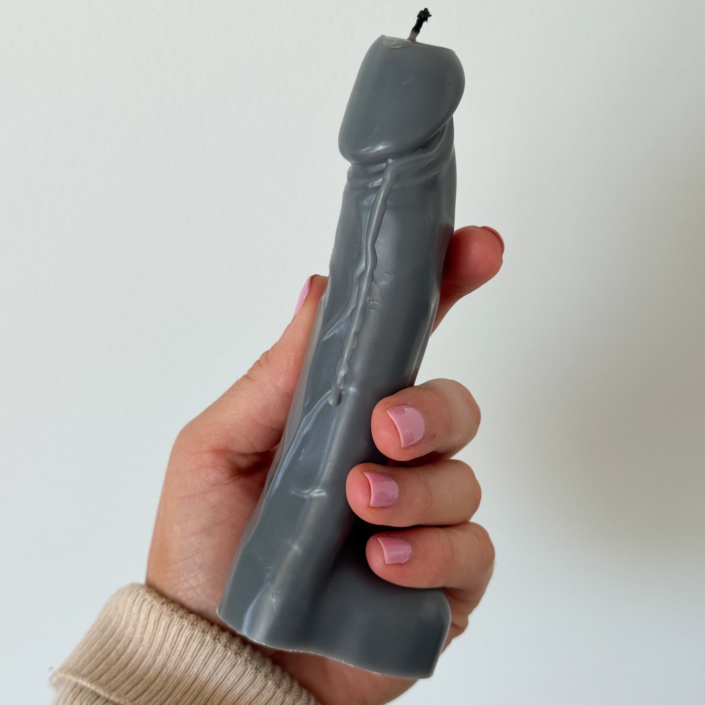 A hand holding a sculptural gray novelty candle shaped like a stylized penis, against a plain off-white background.
