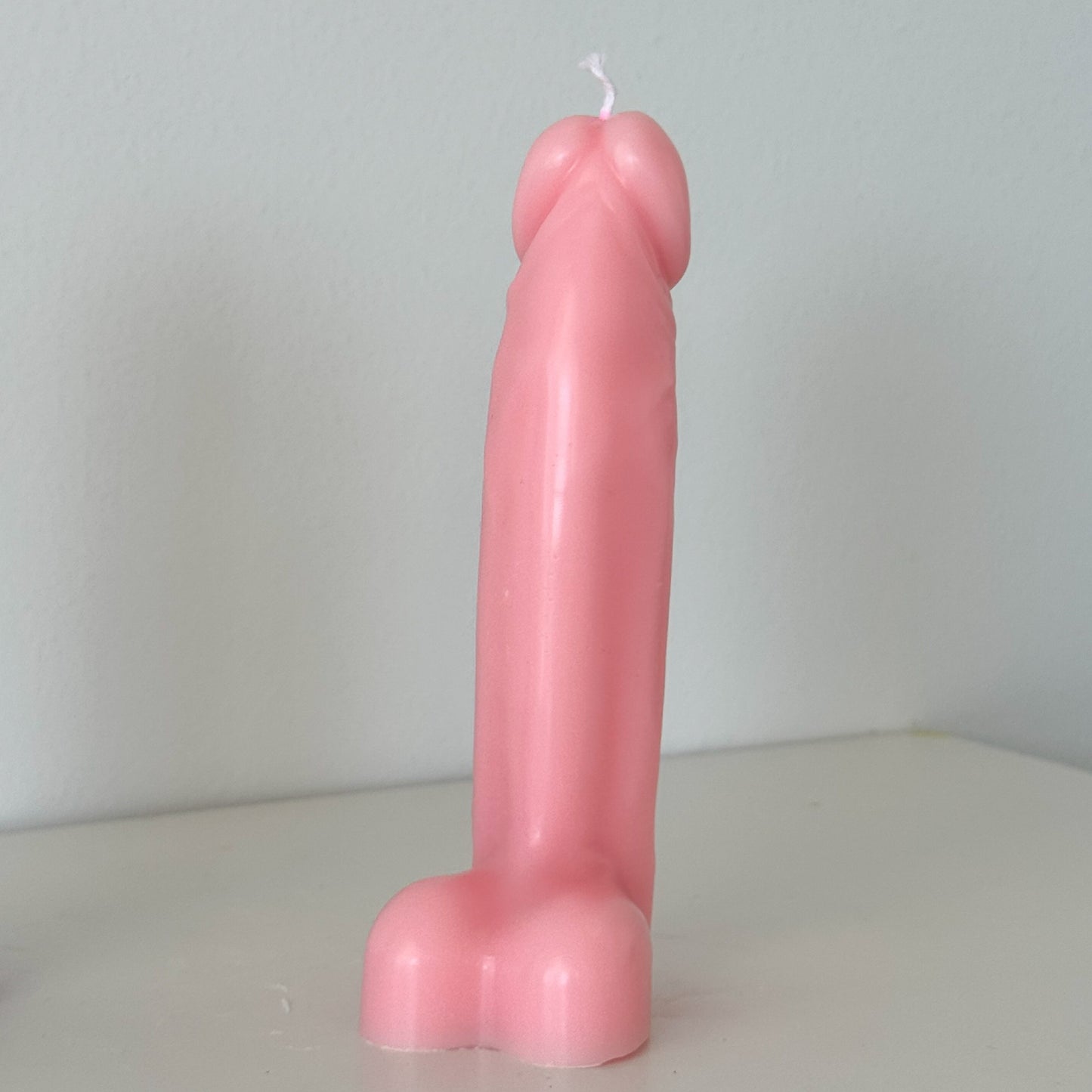 A novelty pink candle shaped like a penis, standing upright on a light-colored surface against a plain light gray wall.