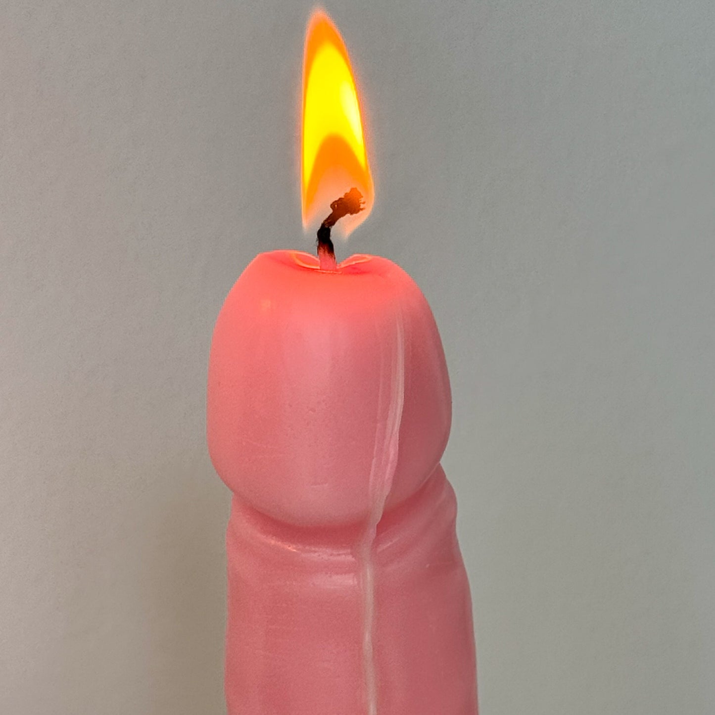 A novelty pink candle shaped like a penis, shown lit with a yellow-orange flame against a plain light gray background.