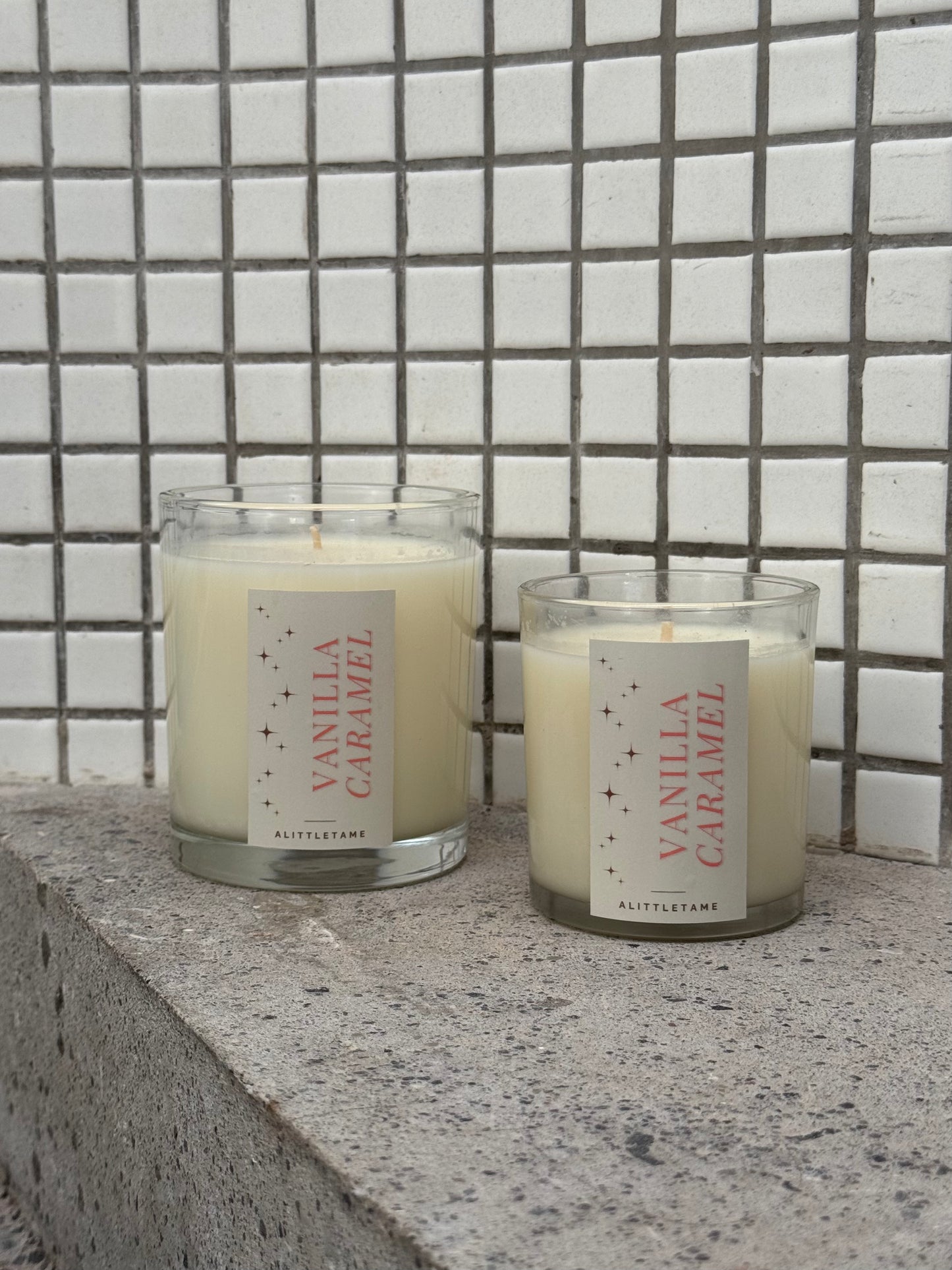 Two vanilla caramel scented candles on a stone surface with a tiled wall background.