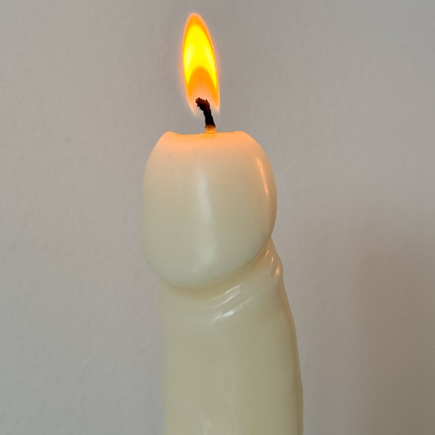 A lit cream-colored pillar candle with a tapered, rounded shape and a small black wick, shown against a plain light gray background.